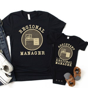 May include: A black t-shirt with the text "Regional Manager" in a circle with a desk and computer icon. A black baby onesie with the text "Assistant to the Regional Manager" in a circle with a desk and computer icon.