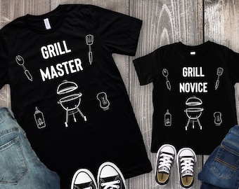 Könnte beinhalten: Zwei schwarze T-Shirts mit weißem Text und Grafiken. Das größere Shirt sagt "Grill Master" und hat einen Grill, einen Spatel, eine Zange und eine Flasche Sauce. Das kleinere Shirt sagt "Grill Novice" und hat die gleichen Grafiken. Beide Shirts werden mit einer blauen Jeans und schwarzen Turnschuhen kombiniert.