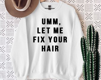 May include: White sweatshirt with the text "Umm, Let Me Fix Your Hair" in black block letters. A brown hat hangs on the wall behind the sweatshirt. A cactus in a white pot sits on a wooden surface.