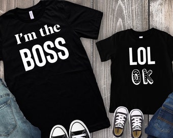 Könnte beinhalten: Zwei schwarze T-Shirts mit weißem Text. Das größere Shirt sagt "I'm the BOSS" und das kleinere Shirt sagt "LOL OK". Die Shirts liegen auf einer Holzoberfläche mit Jeans und schwarzen Turnschuhen.