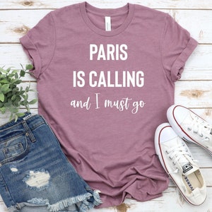 Może przedstawiać: Koszulka w kolorze malwy z napisem "Paris is calling and I must go" w białym kolorze.