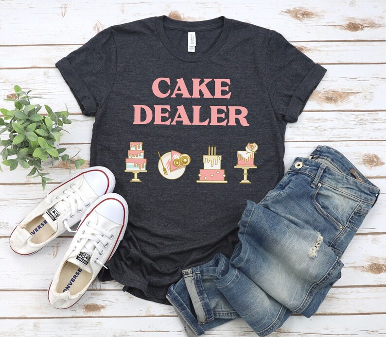 Puede incluir: Una camiseta gris oscuro jaspeada con el texto "CAKE DEALER" en letras rosas. La camiseta presenta una fila de cinco ilustraciones de pasteles coloridos.