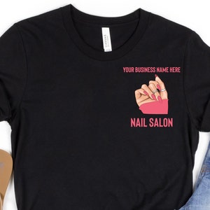 May include: Black t-shirt featuring a nail salon design. The design includes a hand with long, pink nails, wearing rings, and the text "YOUR BUSINESS NAME HERE" and "NAIL SALON" in pink. The shirt is made of a soft material.
