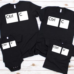 Op de afbeelding: Een set van vier zwarte t-shirts met witte computertoetsenbordtoetsen erop gedrukt. De shirts hebben de toetsen "Ctrl C" en "Ctrl V".