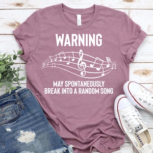 Könnte beinhalten: Ein mauvefarbenes T-Shirt mit dem Text "WARNING MAY SPONTANEOUSLY BREAK INTO A RANDOM SONG" in Weiß. Der Text ist von Noten umgeben.