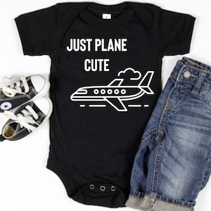 May include: Black bodysuit with white text that reads "Just Plane Cute" and a white airplane graphic. The bodysuit is paired with blue denim shorts and black and white sneakers.
