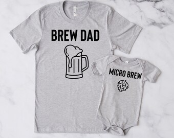 Könnte beinhalten: Ein graues T-Shirt mit dem Text "Brew Dad" und einer Grafik eines Bierkruges. Ein grauer Baby-Strampler mit dem Text "Micro Brew" und einer Grafik einer Hopfenpflanze.