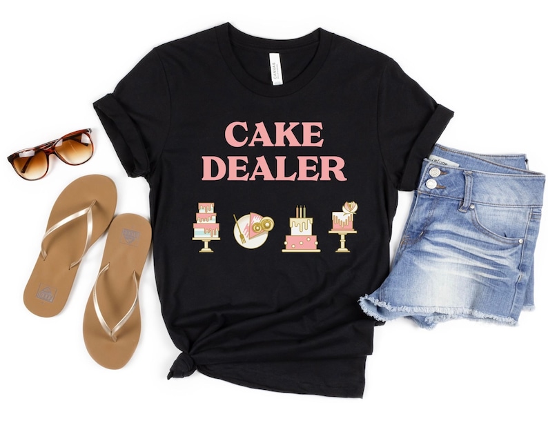 Puede incluir: Camiseta negra con el texto "CAKE DEALER" en letras rosas. La camiseta tiene cuatro peque&ntilde;as im&aacute;genes de pasteles en dorado y rosa.