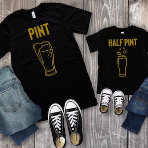 以下が含まれることがあります： 金色のビールグラスのグラフィックとテキストが書かれた黒いTシャツ2枚。大きい方のTシャツには「PINT」、小さい方のTシャツには「HALF PINT」と書いてあります。