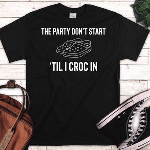 Könnte beinhalten: Schwarzes T-Shirt mit weißem Text, der "The party don't start 'til I croc in." lautet. Das Shirt zeigt ein Motiv mit einem Paar weißen Crocs.