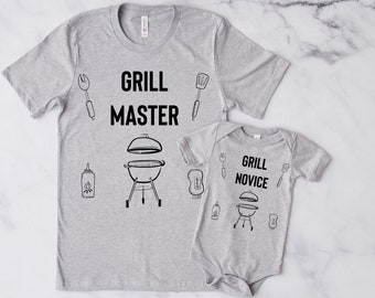 Könnte beinhalten: Zwei graue T-Shirts mit Grill-Motiv. Das größere Shirt trägt den Schriftzug "Grill Master" und zeigt einen Grill, einen Spatel, eine Zange und eine Flasche Sauce. Das kleinere Shirt trägt den Schriftzug "Grill Novice" und zeigt einen Grill, einen Spatel, eine Zange und eine Flasche Sauce.