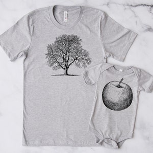 Puede incluir: Una camiseta gris con una ilustración en blanco y negro de un árbol y un body de bebé gris con una ilustración en blanco y negro de una manzana.