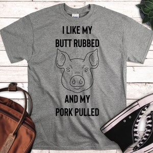 May include: Gray t-shirt with the text "I LIKE MY BUTT RUBBED AND MY PORK PULLED" and a line drawing of a pig's face. The shirt is displayed on a light wood surface with a brown bag and black high-top sneakers.