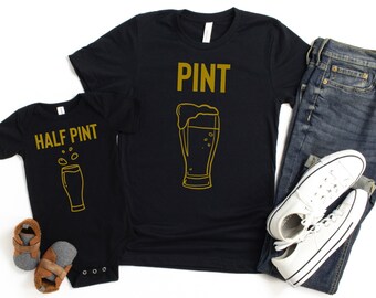 May include: A black t-shirt with the word "PINT" in gold lettering and a gold outline of a beer glass. A black baby onesie with the words "HALF PINT" in gold lettering and a gold outline of a smaller beer glass.