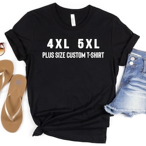 May include: Black t-shirt with white text that reads "4XL 5XL PLUS SIZE CUSTOM T-SHIRT". The shirt is laid flat on a white background with a pair of brown flip flops and denim shorts.