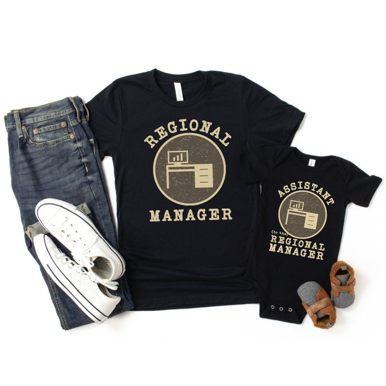 May include: A black t-shirt with the text "Regional Manager" in a circle with a desk and computer icon. A black baby onesie with the text "Assistant to the Regional Manager" in a circle with a desk and computer icon.