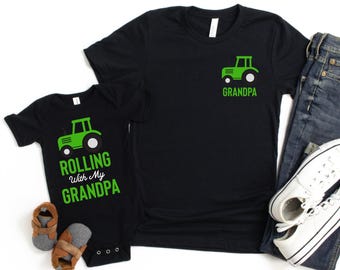 May include: Black t-shirts and a baby onesie with green tractor graphics. The adult shirt has a tractor graphic and the word "Grandpa". The onesie says "Rolling With My Grandpa". Blue jeans and white sneakers are also in the image.