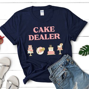 Puede incluir: Una camiseta azul marino con el texto "CAKE DEALER" en letras rosas. Hay cinco peque&ntilde;as ilustraciones de pasteles en dorado y rosa en la camiseta.