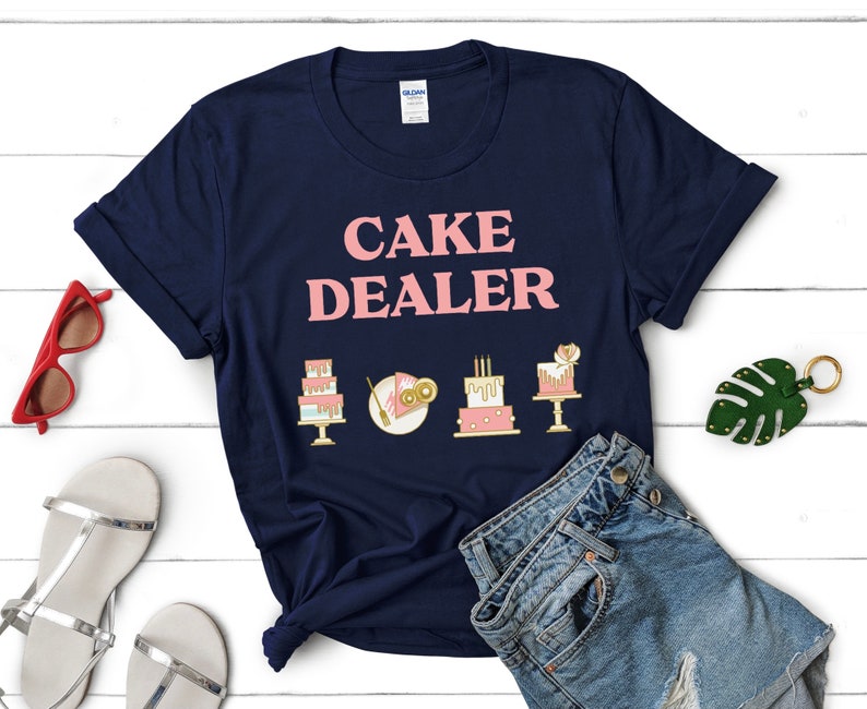 Puede incluir: Una camiseta azul marino con el texto "CAKE DEALER" en letras rosas. Hay cinco peque&ntilde;as ilustraciones de pasteles en dorado y rosa en la camiseta.