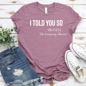May include: A mauve t-shirt with the text "I told you so, Signed, "The Conspiracy Theorist" printed in white.