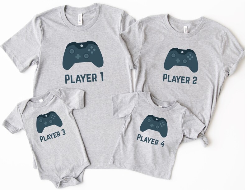May include: Four gray t-shirts and a onesie with a video game controller graphic. Each shirt is labeled "Player 1", "Player 2", "Player 3", and "Player 4". The controller is dark blue.