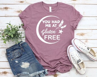 Könnte beinhalten: Ein mauvefarbenes T-Shirt mit dem Text "You had me at gluten free" in einem weißen Kreis mit einem Halbmond und einem Stern. Das Shirt liegt auf einer weißen Holzoberfläche mit einem Paar weißen Canvas-Schuhen und einer blauen Jeans-Shorts.