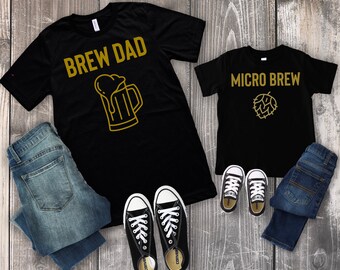 Könnte beinhalten: Zwei schwarze T-Shirts mit goldfarbenem Text und Grafiken. Das größere Shirt trägt die Aufschrift "Brew Dad" mit einer Bierkrüg-Grafik. Das kleinere Shirt trägt die Aufschrift "Micro Brew" mit einer Hopfengrafik. Die Shirts liegen auf einer Holzoberfläche mit Jeans und Turnschuhen.
