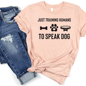 Pode incluir: Uma camiseta rosa claro com o texto "JUST TRAINING HUMANS TO SPEAK DOG" em preto. O texto é acompanhado por um osso, uma pegada de pata e uma tigela para cães. A camiseta é combinada com jeans azul.