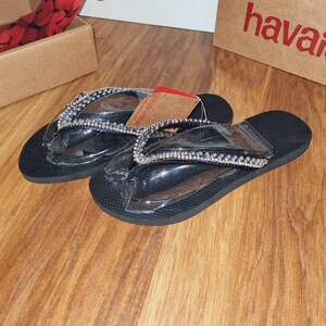 May include: A pair of black flip-flops with clear straps and silver rhinestones. The flip-flops are on a wooden surface.