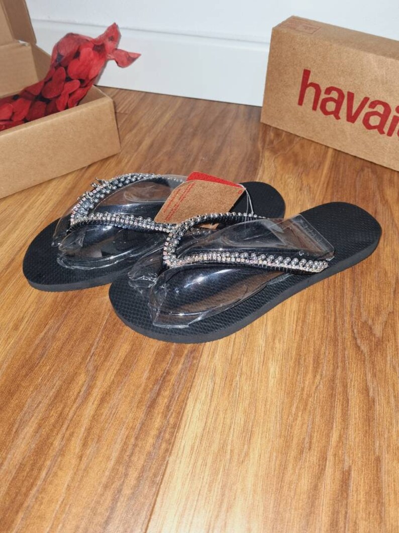 May include: A pair of black flip-flops with clear straps and silver rhinestones. The flip-flops are on a wooden surface.
