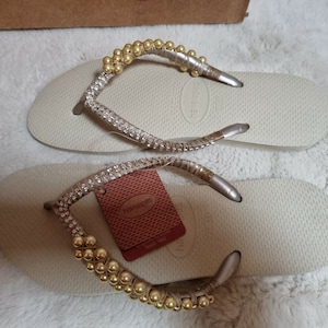 May include: A pair of beige flip-flops with gold and silver embellishments. The flip-flops have a rhinestones strap and a gold beaded strap. The brand name "havaianas" is visible on the sole of the flip-flop.