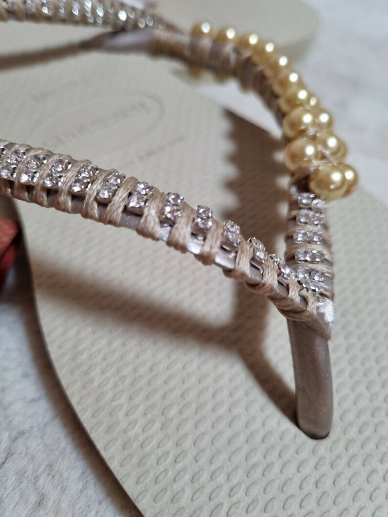 May include: Beige flip-flops with a decorative strap adorned with small rhinestones and pearl-like beads.