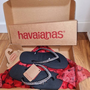 May include: Black flip-flops with a silver rhinestone embellishment, in a cardboard box with the Havaianas logo. The flip-flops are on a red and white patterned tissue paper.