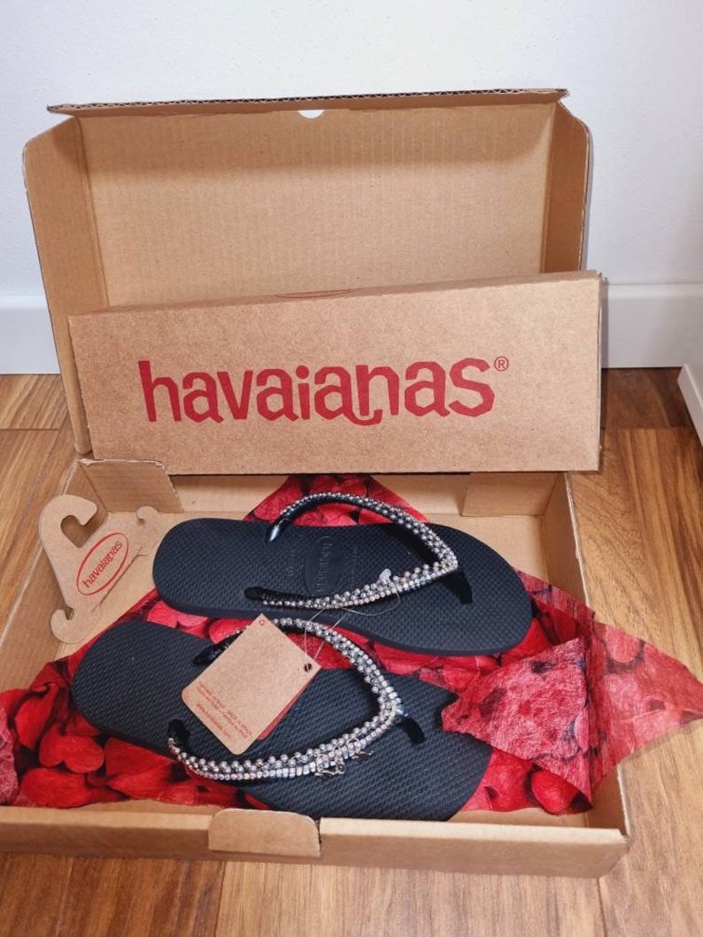 May include: Black flip-flops with a silver rhinestone embellishment, in a cardboard box with the Havaianas logo. The flip-flops are on a red and white patterned tissue paper.