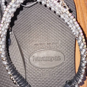 May include: Black flip-flops with a silver and black beaded strap. The flip-flops have the text "havaianas" and "37-38" printed on the sole. The size is also printed as "USA 7/8W EUR 39/40".