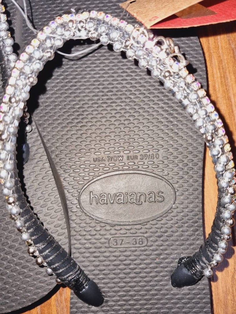 May include: Black flip-flops with a silver and black beaded strap. The flip-flops have the text "havaianas" and "37-38" printed on the sole. The size is also printed as "USA 7/8W EUR 39/40".