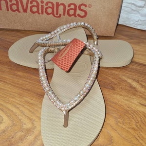 Op de afbeelding: Beige flip-flops met een gouden en parelversiering. De flip-flops liggen op een houten oppervlak. De merknaam "havaianas" is zichtbaar op een doos op de achtergrond.