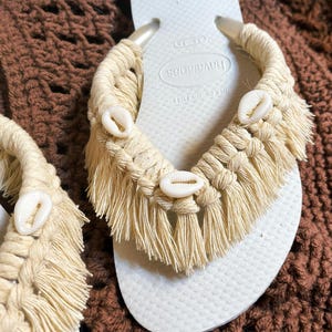 May include: White Havaianas flip-flops with a macrame design in natural beige. The macrame features fringe and is adorned with small, white seashells. A stylish and beachy footwear option.
