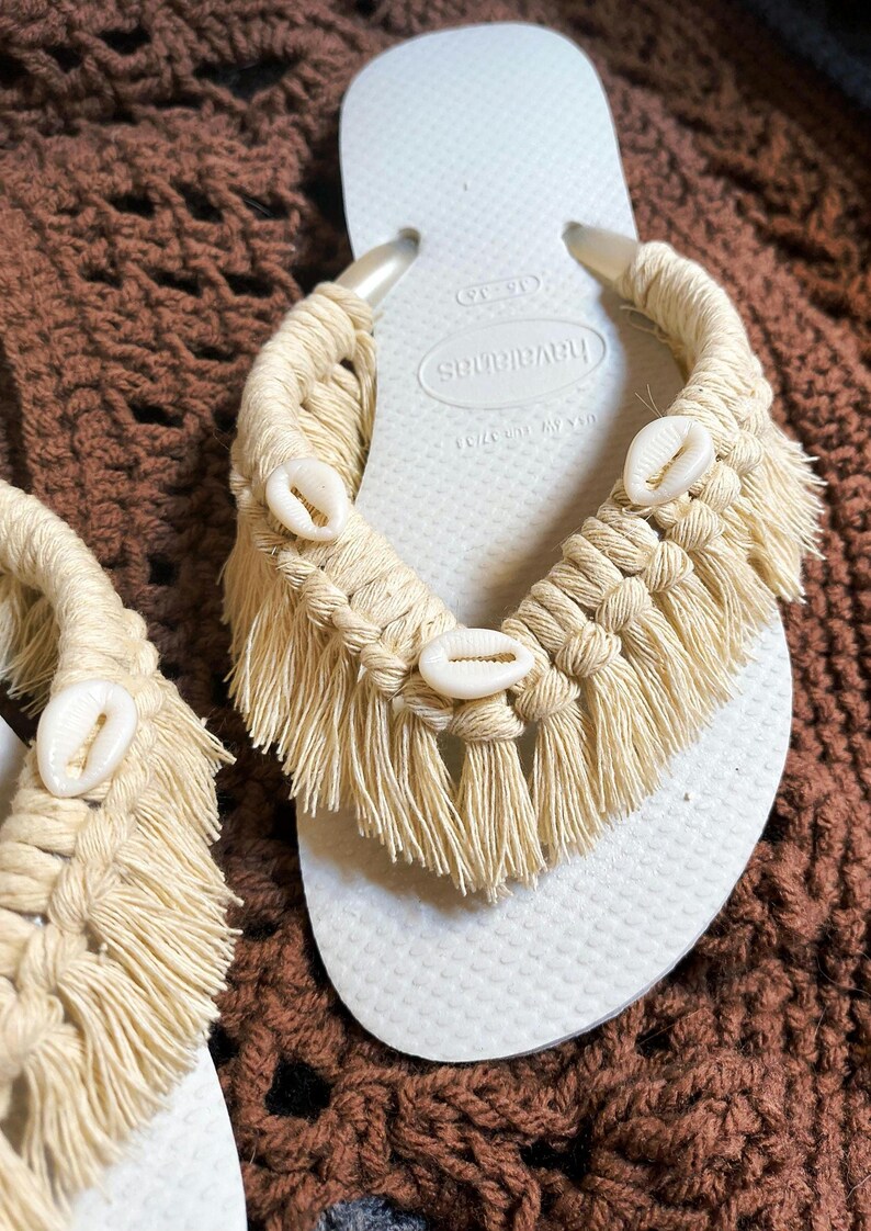 May include: White Havaianas flip-flops with a macrame design in natural beige. The macrame features fringe and is adorned with small, white seashells. A stylish and beachy footwear option.