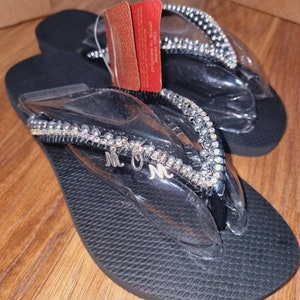 May include: Black flip flops with a silver rhinestone trim and the letters "MOM" in silver on the strap.