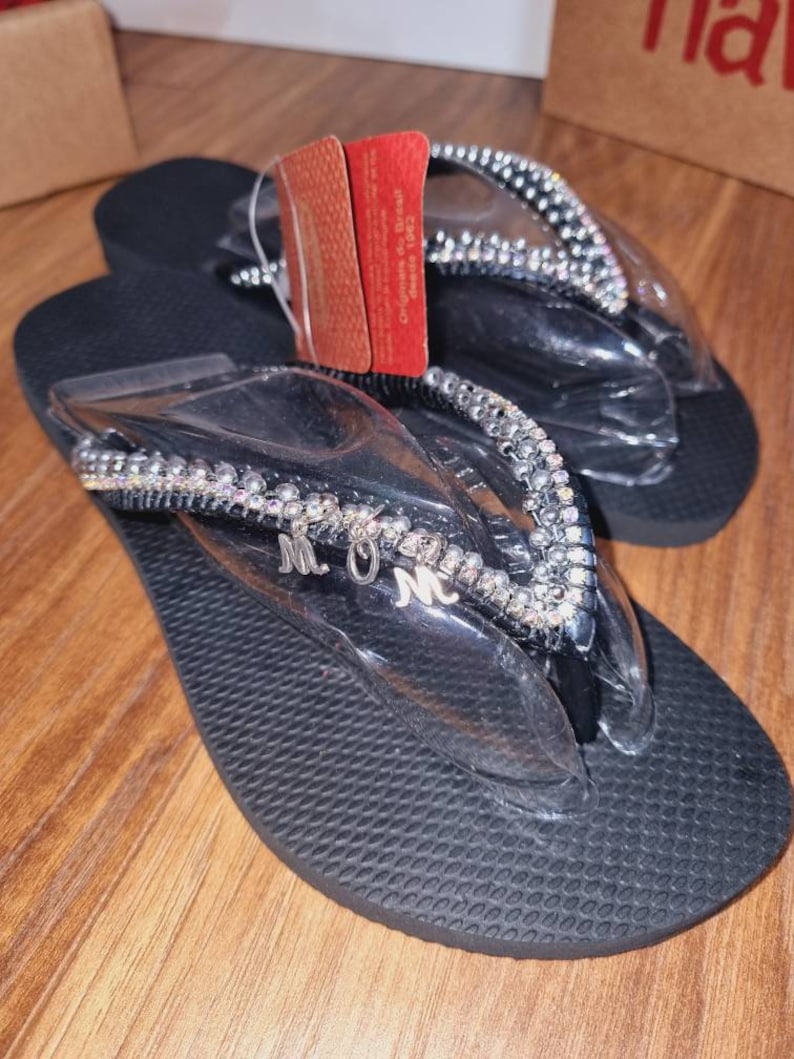 May include: Black flip flops with a silver rhinestone trim and the letters "MOM" in silver on the strap.