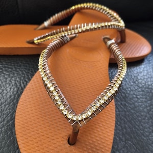 May include: A pair of brown flip-flops with a gold and clear rhinestone embellishment on the strap.