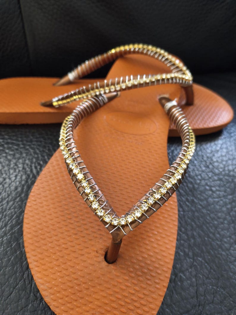 May include: A pair of brown flip-flops with a gold and clear rhinestone embellishment on the strap.
