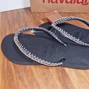 May include: Black flip-flops with a silver rhinestone strap. The strap is decorated with small, round rhinestones.