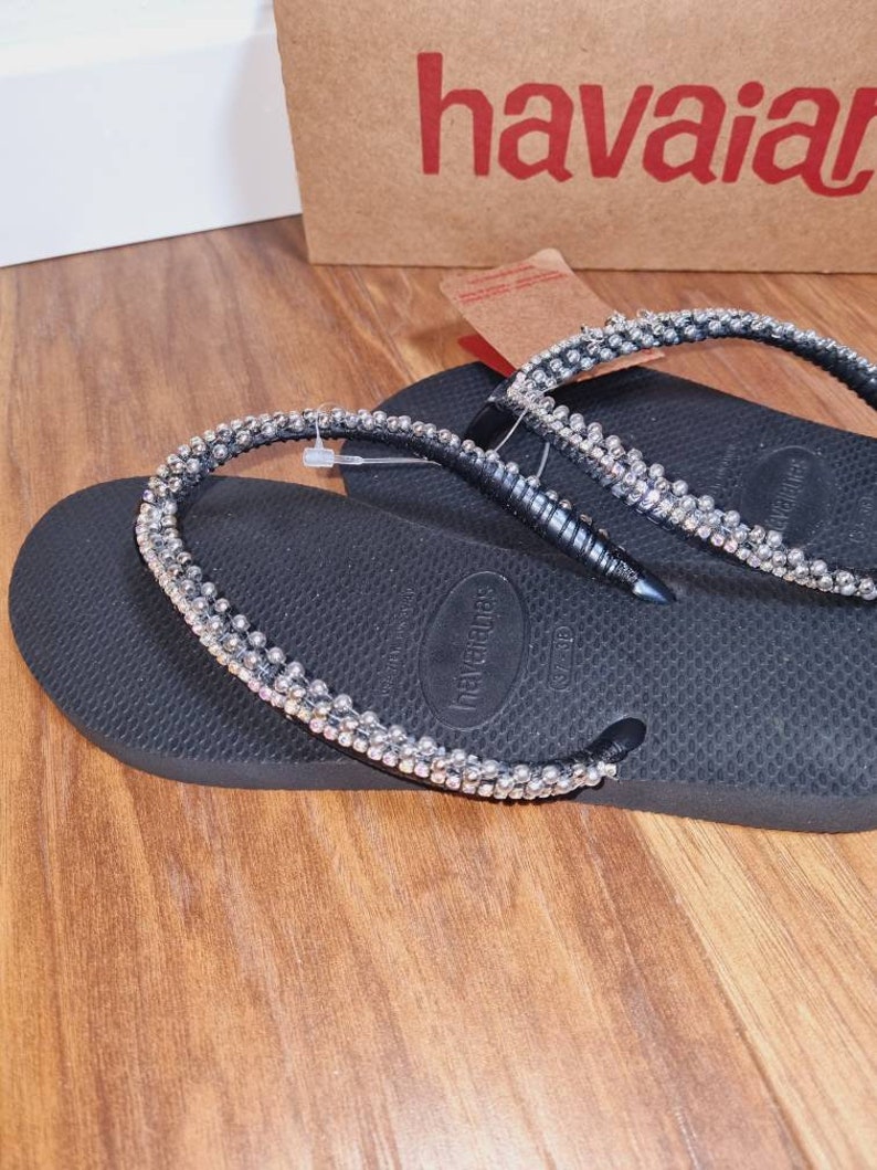 May include: Black flip-flops with a silver rhinestone strap. The strap is decorated with small, round rhinestones.