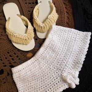 May include: A pair of off-white flip-flops with beige macrame straps adorned with seashells. A white crochet top is also visible. The items are displayed on a brown, textured surface, suggesting a beach or summer theme.