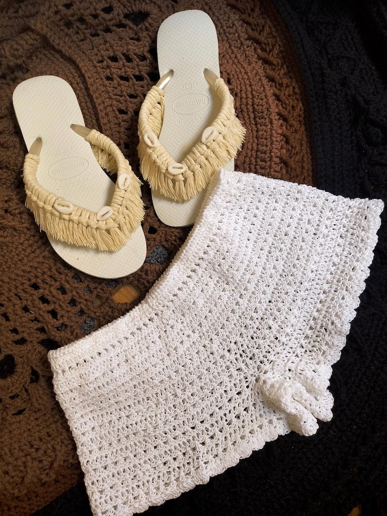 May include: A pair of off-white flip-flops with beige macrame straps adorned with seashells. A white crochet top is also visible. The items are displayed on a brown, textured surface, suggesting a beach or summer theme.