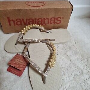 May include: A pair of beige Havaianas flip-flops with a gold and silver beaded strap. The flip-flops are on a white surface.