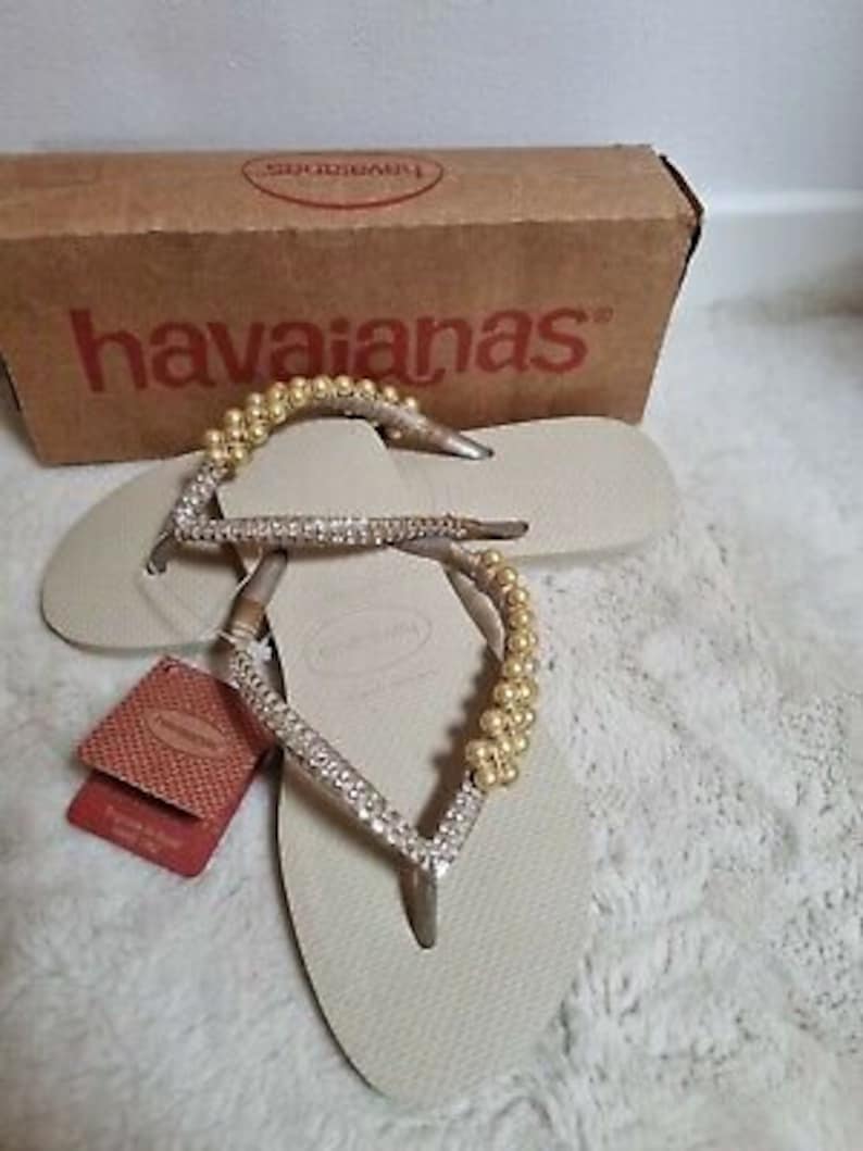 May include: A pair of beige Havaianas flip-flops with a gold and silver beaded strap. The flip-flops are on a white surface.