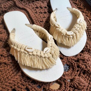 May include: A pair of off-white flip-flops with macrame detailing. The straps are adorned with beige fringe and small seashells. The flip-flops are displayed on a brown crocheted surface.